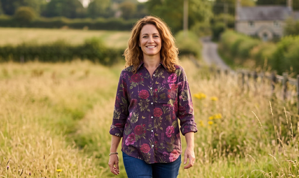 A woman walking through a grassy field wearing a purple floral button-up shirt, blue jeans, and brown knee-high boots.