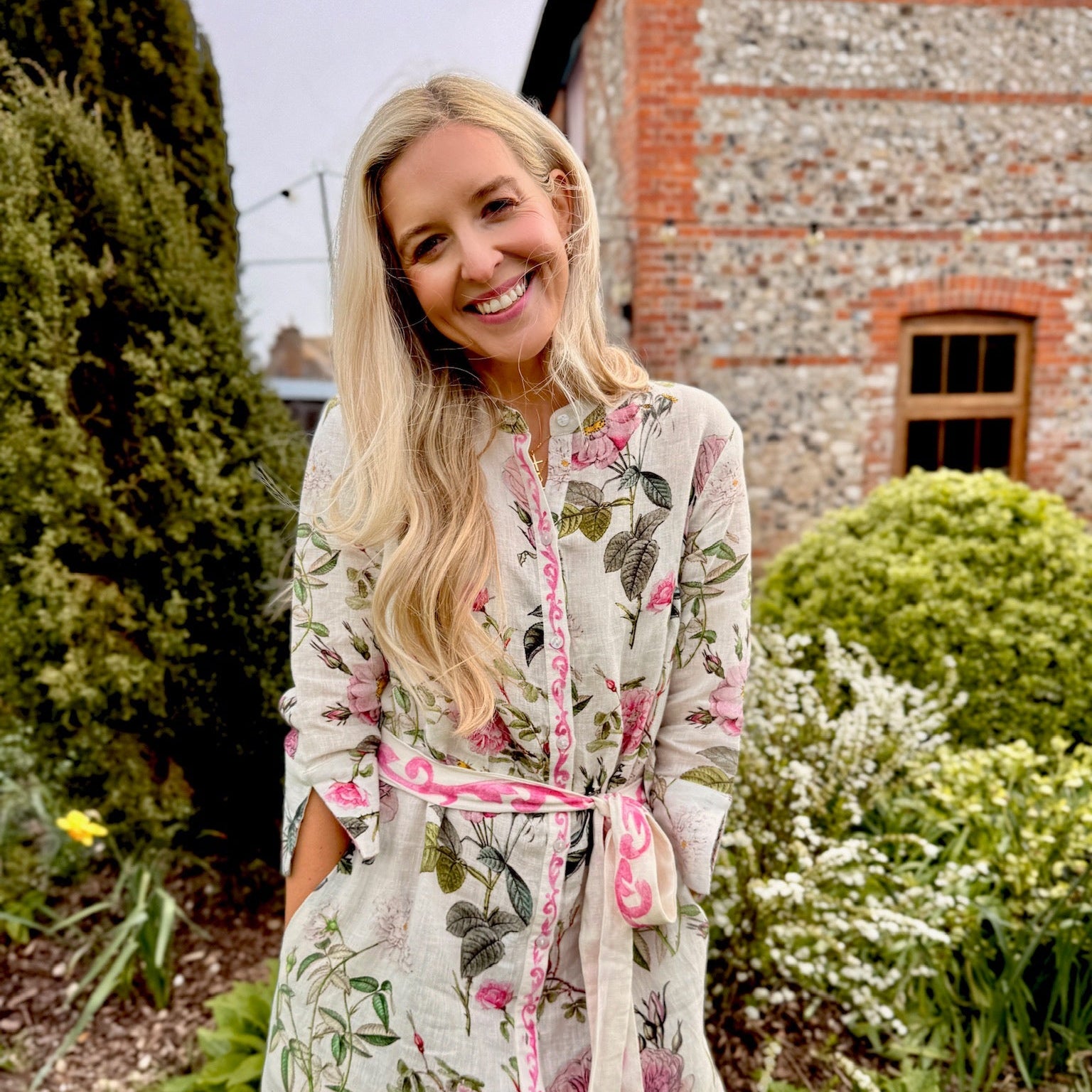 Woman wearing a floral dress standing outdoors with a brick building and greenery in the background