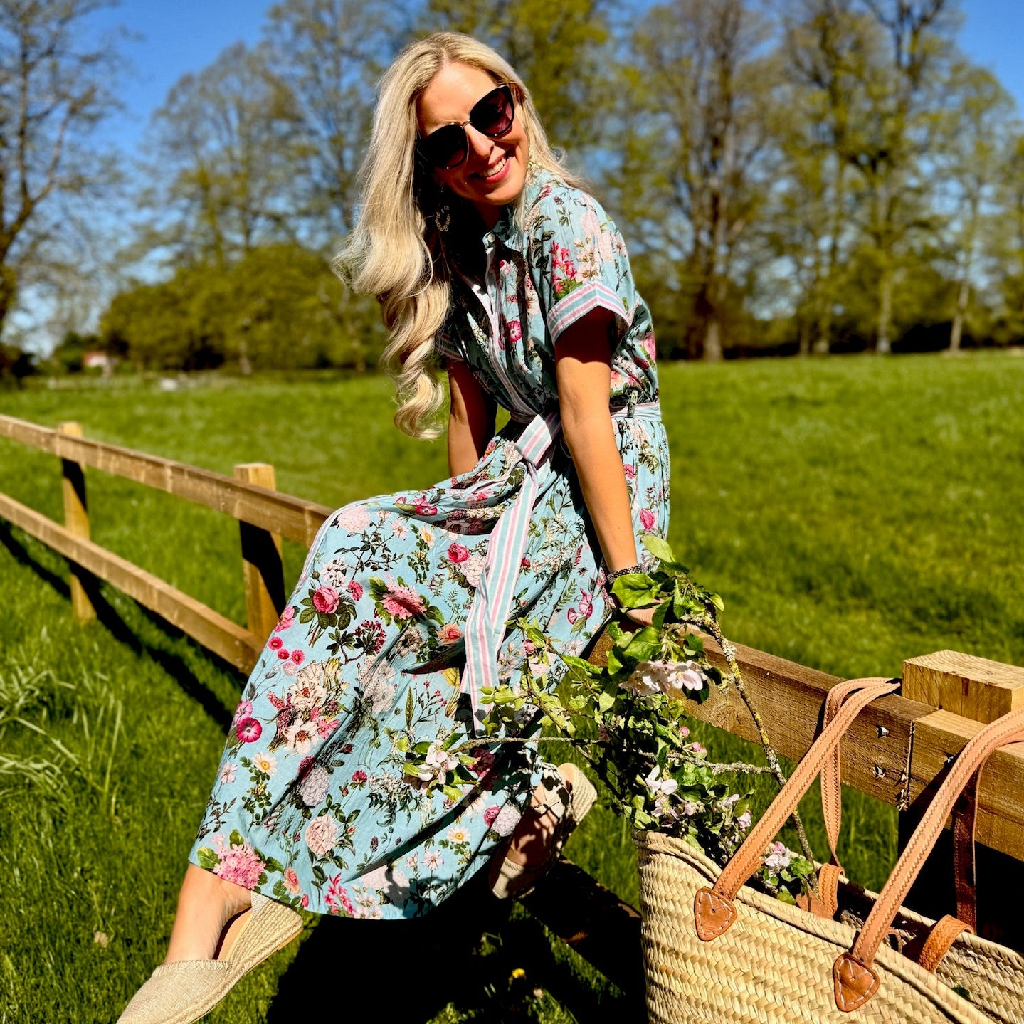 Woman in a Rosehip floral dress sitting on a wooden fence in a field with a straw bag beside her.