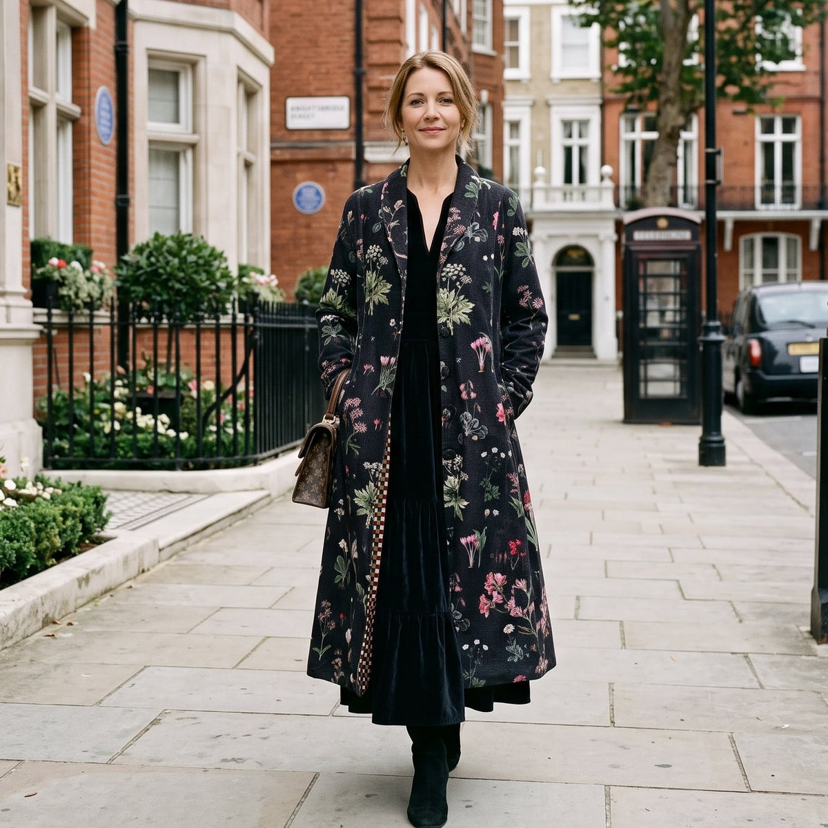 A woman stands on a city sidewalk wearing a long black floral coat over a black dress, with brick buildings, street lamps, and parked cars in the background.
