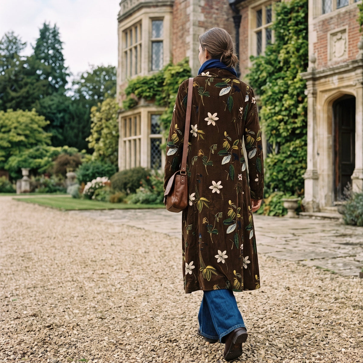 A woman walks away on a gravel path toward a large historic house, wearing a long Rosehip Carla Coat in Enchanted Wood with blue wide-leg pants, and carrying a brown shoulder bag.