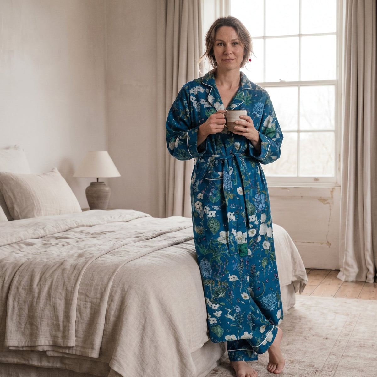 A woman wearing a Rosehip Cool Nights dressing gown over her pyjamas, She stands barefoot in a softly lit bedroom with a bed, curtains, and a fireplace visible in the background.