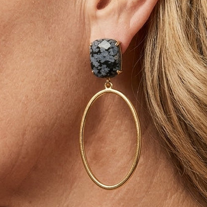 A close-up side profile of a woman with blonde hair wearing a gold hoop earring with a textured dark gray stone stud, along with tortoiseshell eyeglasses.