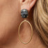 A close-up side profile of a woman with blonde hair wearing a gold hoop earring with a textured dark gray stone stud, along with tortoiseshell eyeglasses.