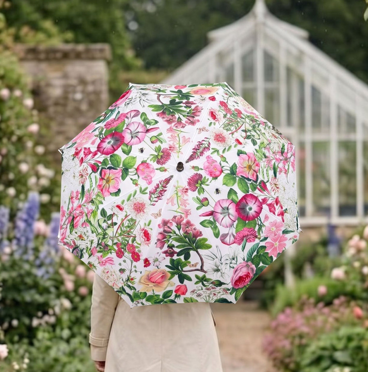 A person walking down a garden path holding a white umbrella with a vibrant pink and green floral print. The scene is set in a lush garden with pink roses, purple flowers, and a glass greenhouse in the background.
