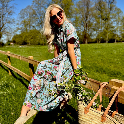 Woman in a Rosehip floral dress sitting on a wooden fence in a field with a straw bag beside her.