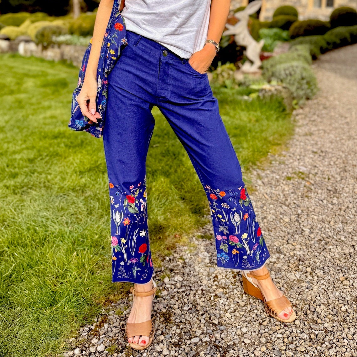 Woman in blue floral Rosehip Trousers and white shirt standing on a gravel path with a house in the background