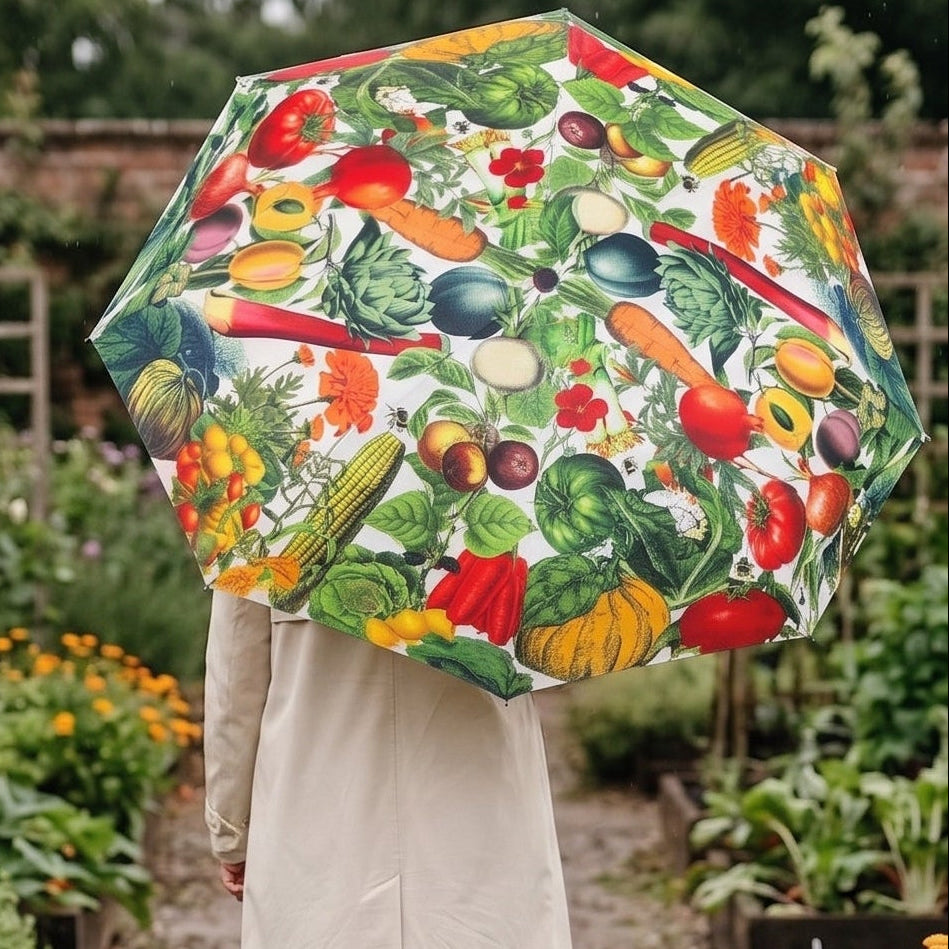 A person standing in a garden holding a large open umbrella printed with colorful vegetables on a white background. The person is wearing a light beige coat or dress.