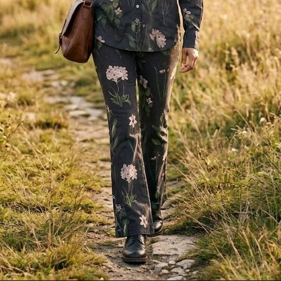 A woman walking on a dirt path through a grassy field at sunset, wearing a dark floral button-up shirt and matching pants, with a brown shoulder bag.