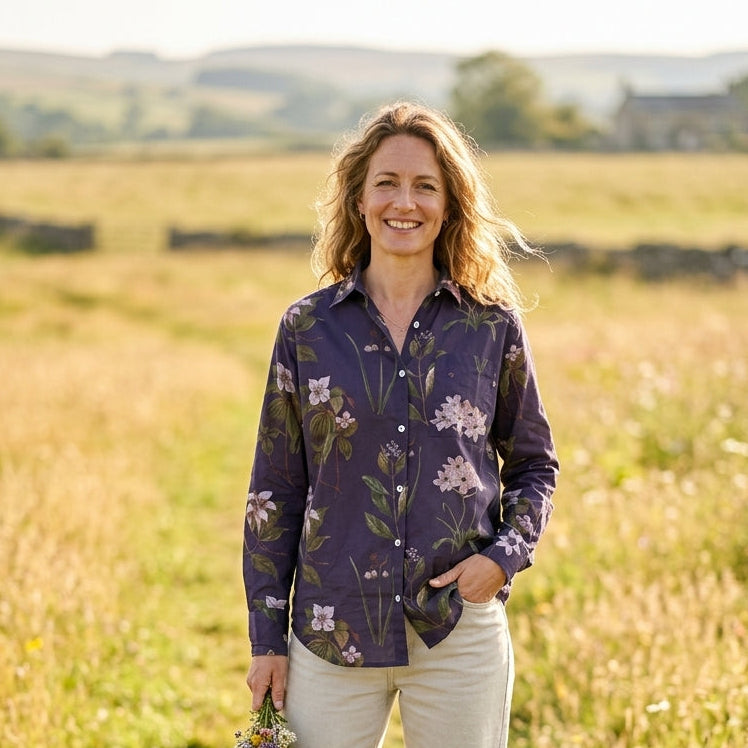 A woman stands in a sunlit grassy field wearing a dark purple floral button-up shirt and light beige trousers, holding a small bouquet of wildflowers.