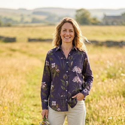 A woman stands in a sunlit grassy field wearing a dark purple floral button-up shirt and light beige trousers, holding a small bouquet of wildflowers.