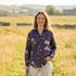 A woman stands in a sunlit grassy field wearing a dark purple floral button-up shirt and light beige trousers, holding a small bouquet of wildflowers.
