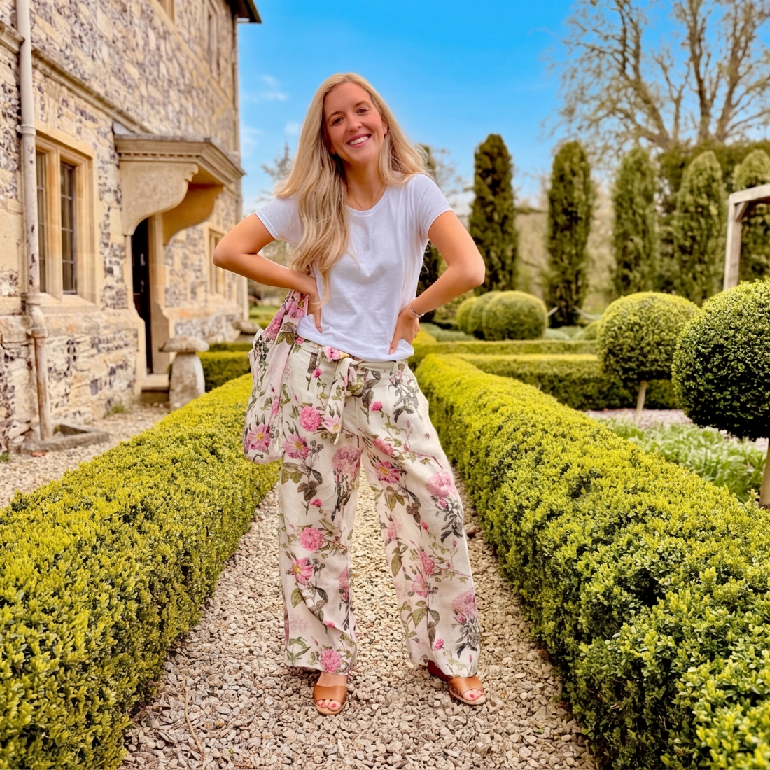 Woman wearing Rosehip Tessa Linen trousers standing in a garden with manicured hedges and a building in the background